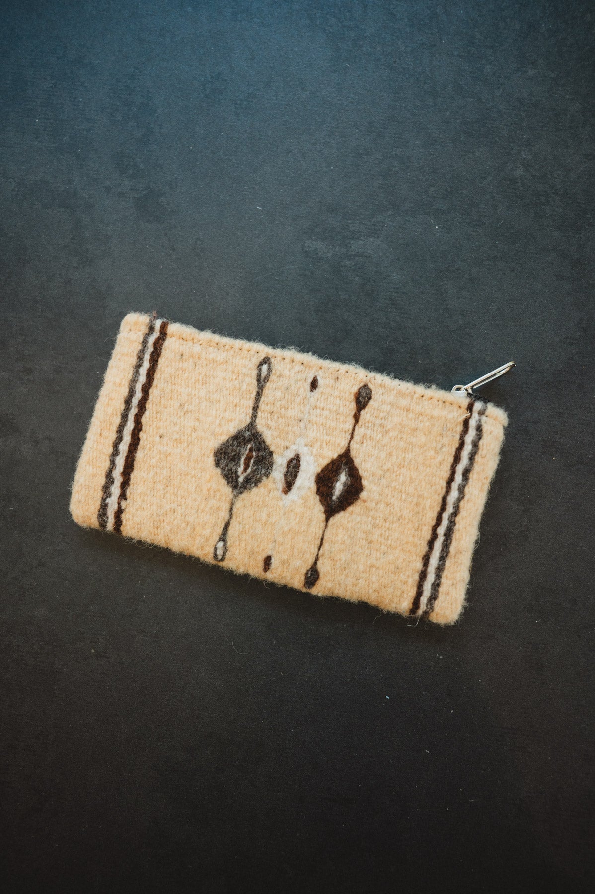 A small woven zippered pouch or wallet in a light beige wool-like material with dark brown geometric and vertical stripe patterns, photographed on a dark textured background.