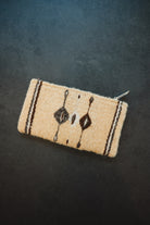 A small woven zippered pouch or wallet in a light beige wool-like material with dark brown geometric and vertical stripe patterns, photographed on a dark textured background.