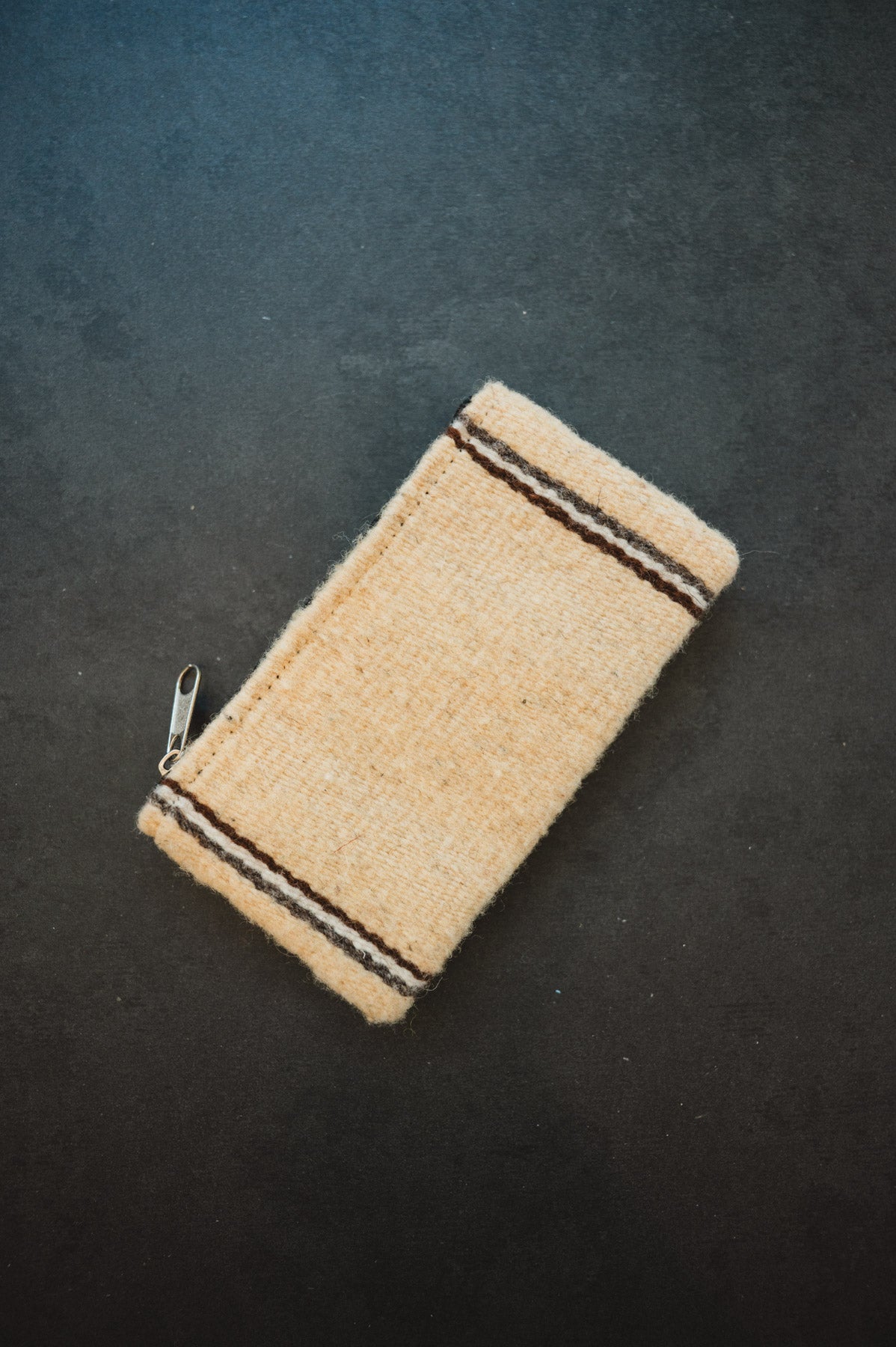 A small woven zippered pouch or case in a natural beige material, featuring two dark brown stripes near the top and bottom edges, photographed on a dark textured background.
