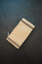 A small woven zippered pouch or case in a natural beige material, featuring two dark brown stripes near the top and bottom edges, photographed on a dark textured background.