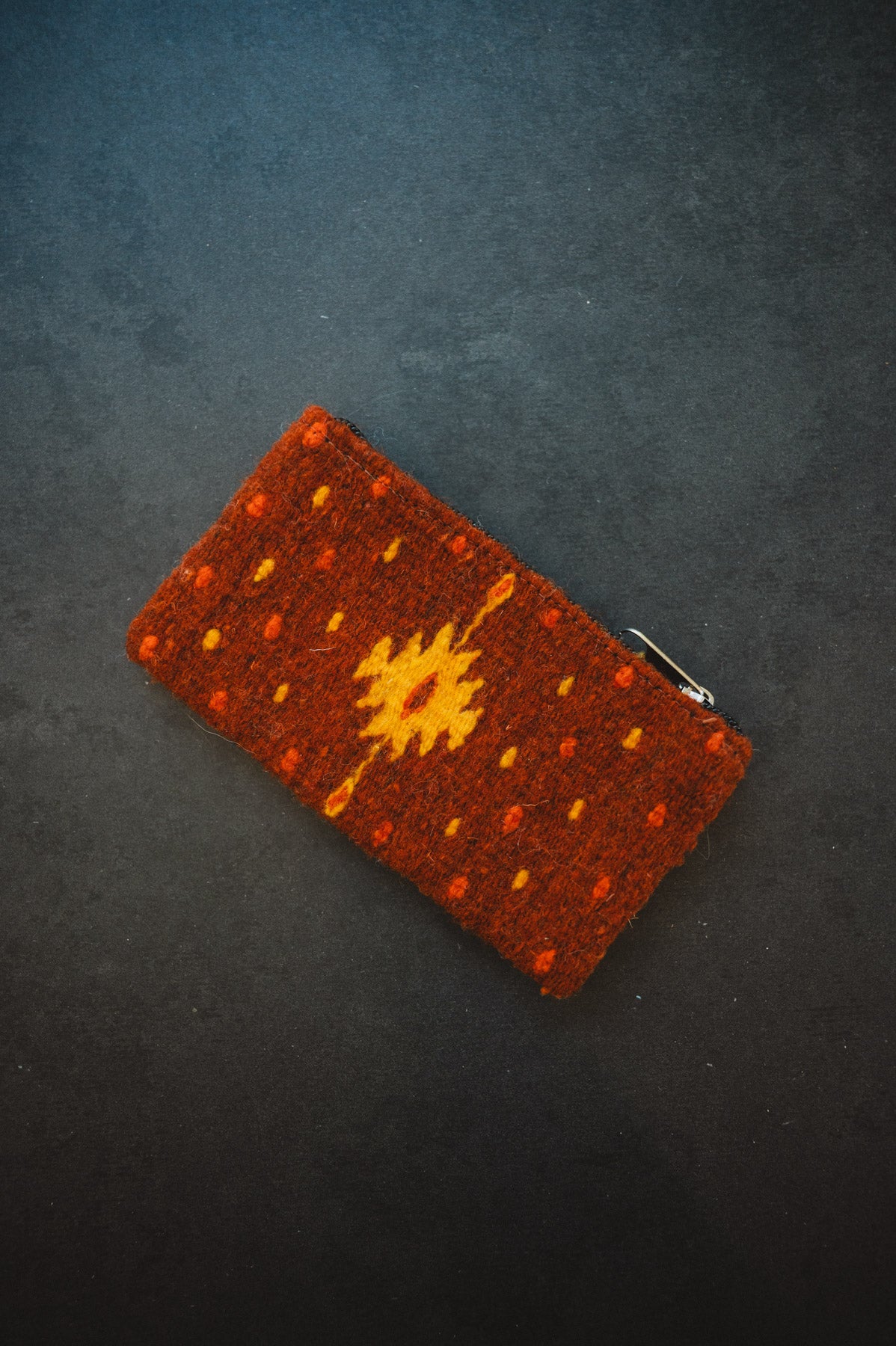 A small rectangular woven pouch or clutch in rust brown with a yellow geometric sun motif and orange dotted accents, photographed on a dark textured background.
