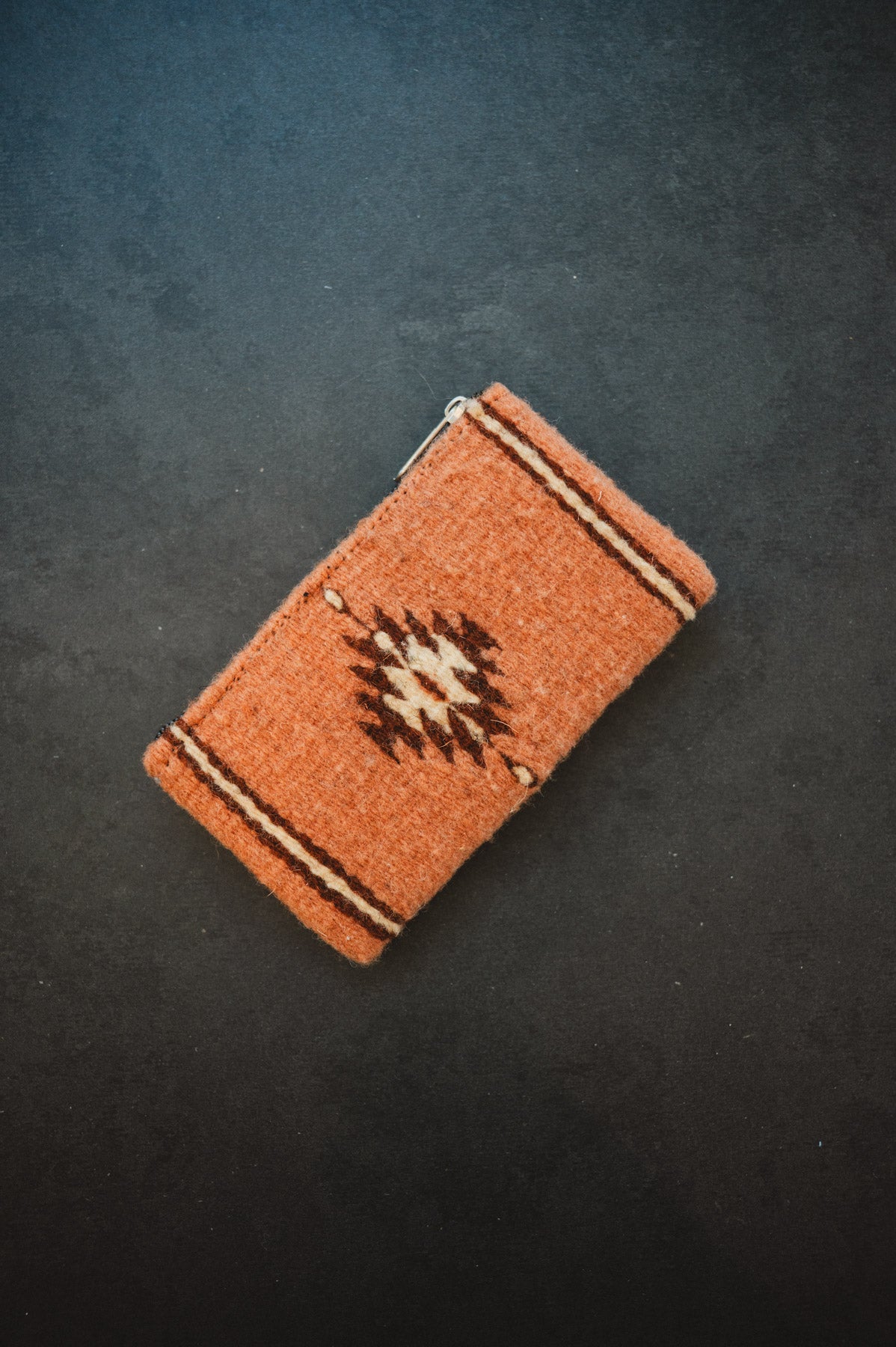 A small woven zippered pouch or wallet in terracotta orange with a Southwestern-style geometric pattern in black, beige, and brown, photographed on a dark textured background.