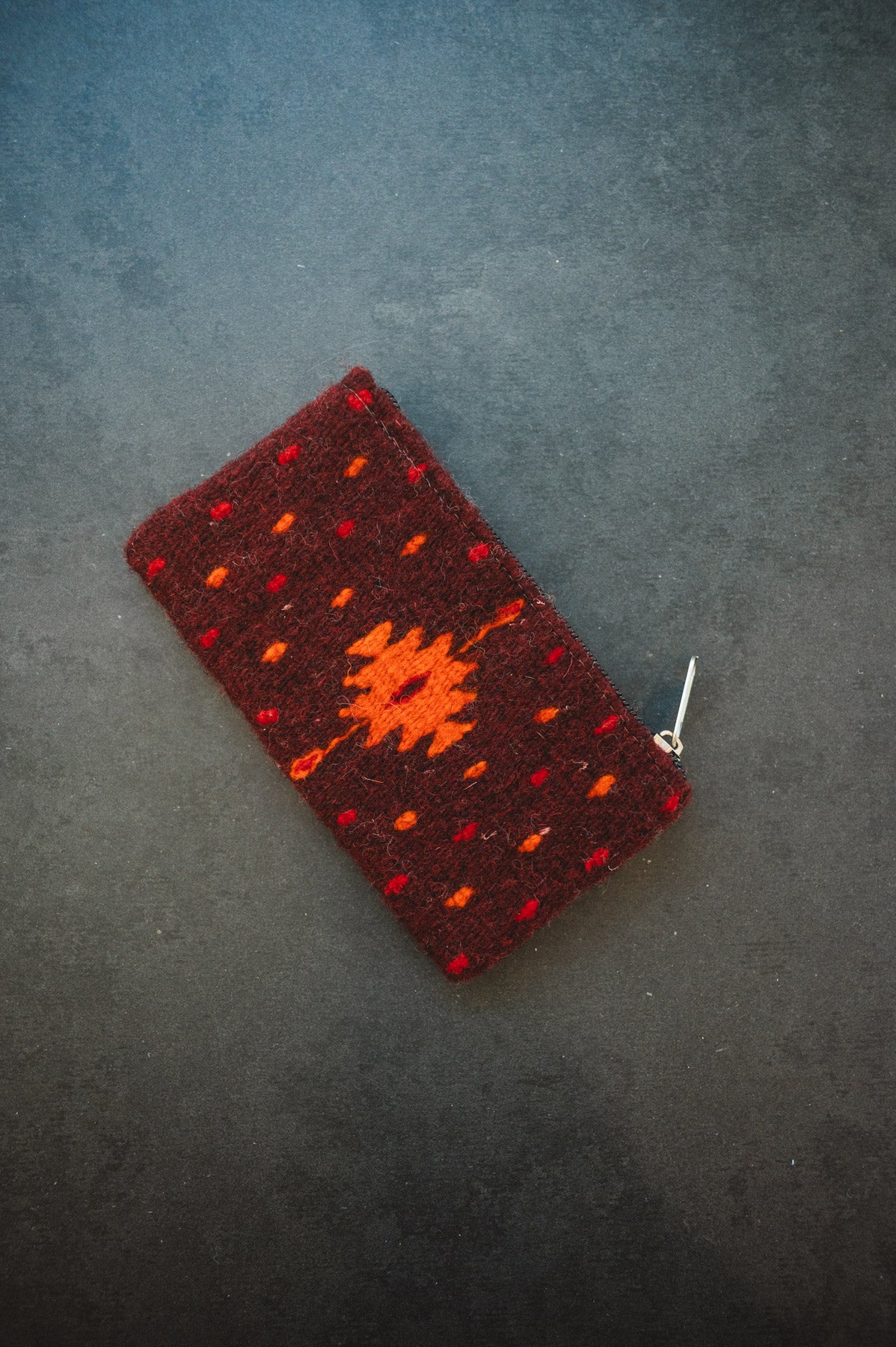 A small woven zippered pouch or wallet in dark red with orange geometric patterns, photographed on a dark textured surface.