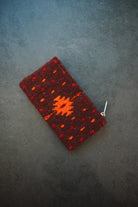 A small woven zippered pouch or wallet in dark red with orange geometric patterns, photographed on a dark textured surface.