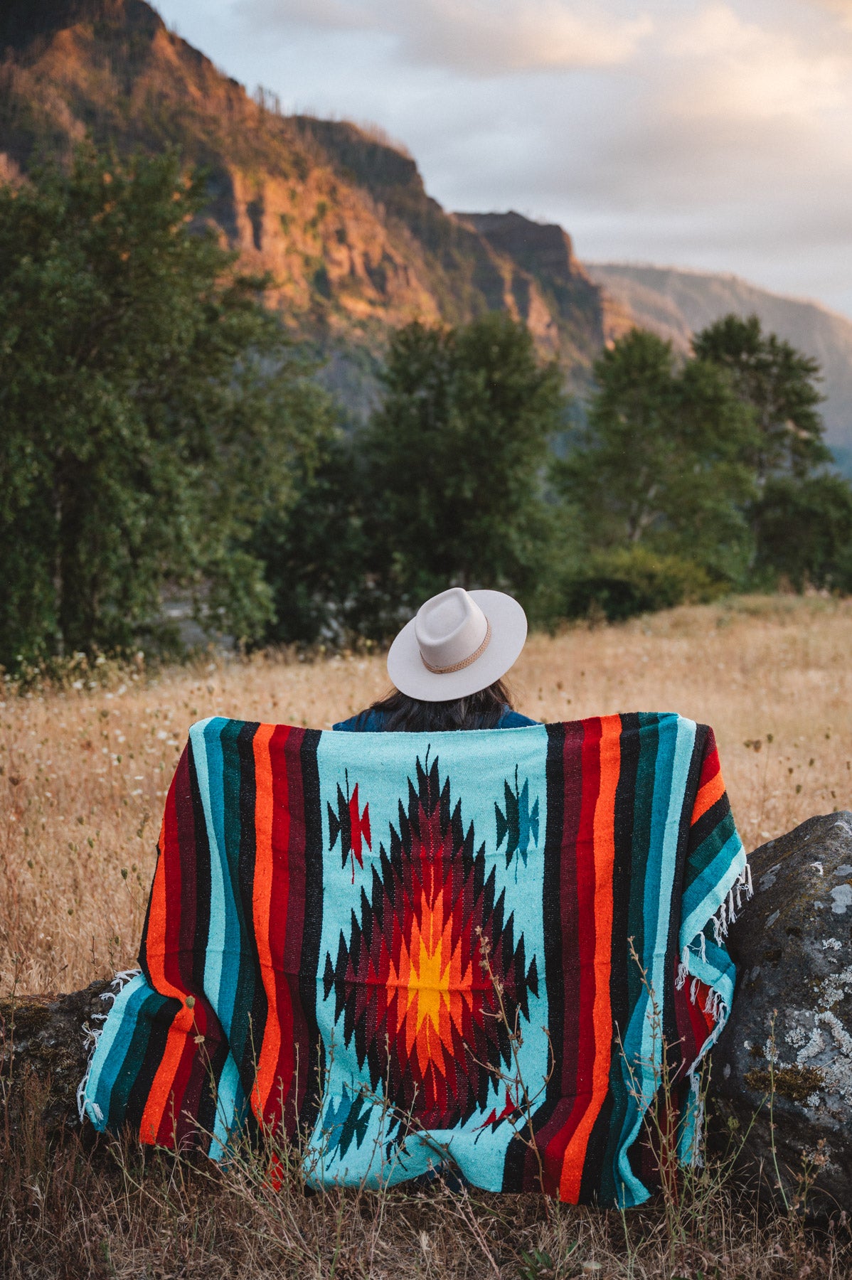 A person sitting outdoors in a grassy field with mountains and trees in the background, viewed from behind while wearing a white wide-brim hat and wrapped in a colorful Southwestern-style woven blanket with turquoise, red, orange, black, and cream geometric patterns.