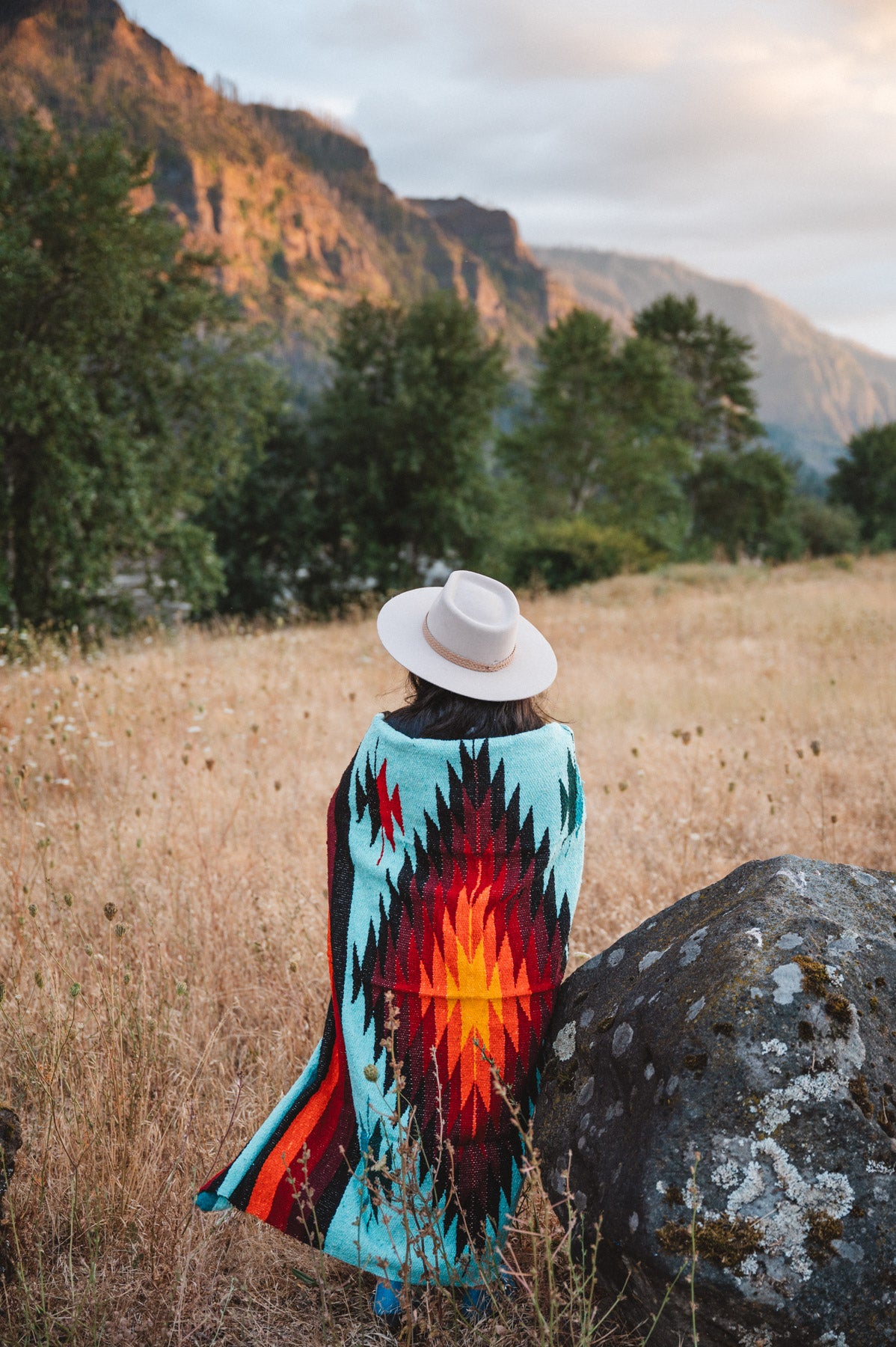 A person seen from behind sitting in a grassy field with mountains and trees in the background, wearing a white hat and a colorful southwestern-style blanket or poncho with a turquoise, red, orange, and black geometric pattern.