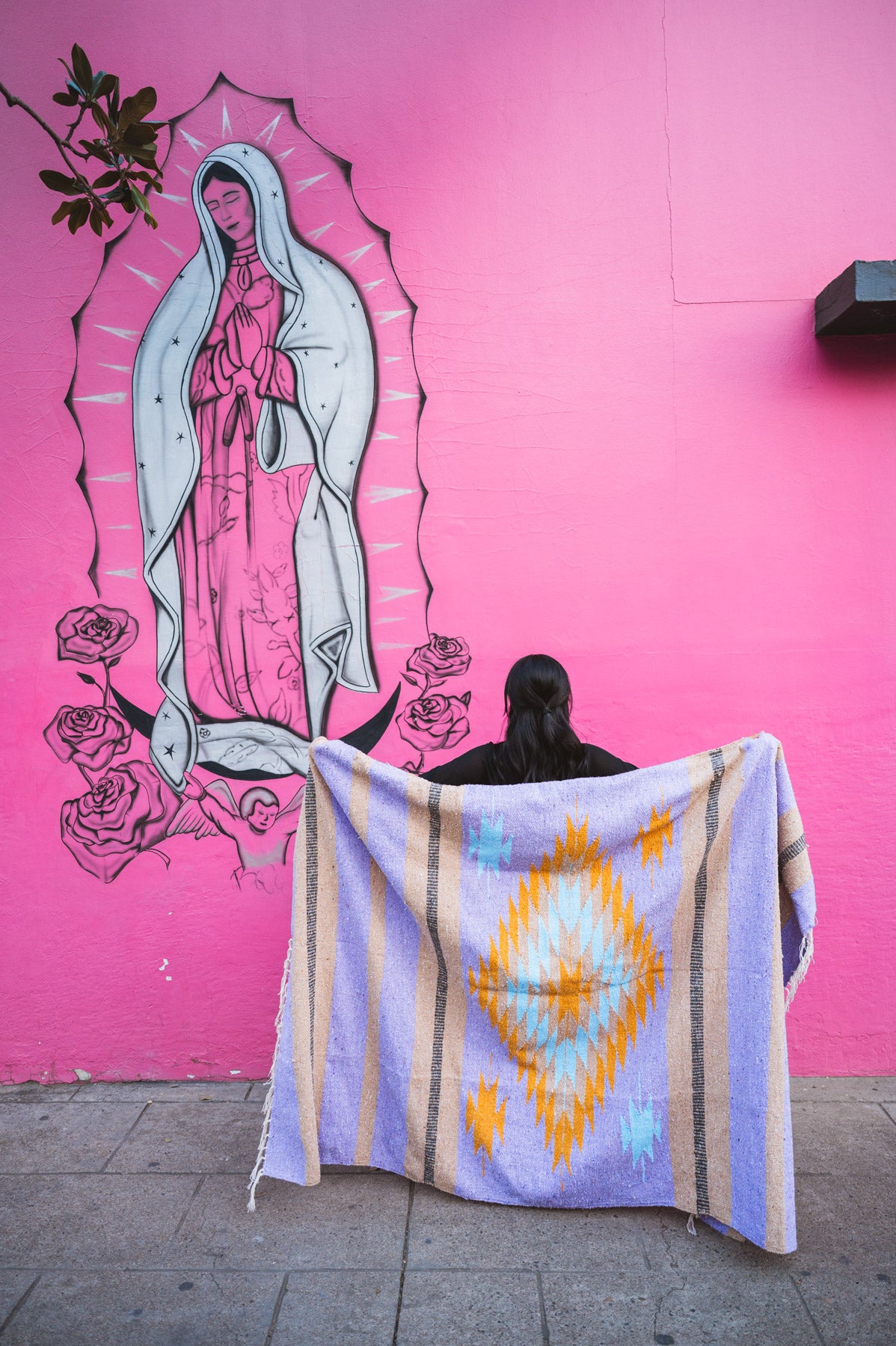 Person holding a colorful blanket in front of a pink wall with a mural of Our Lady of Guadalupe.