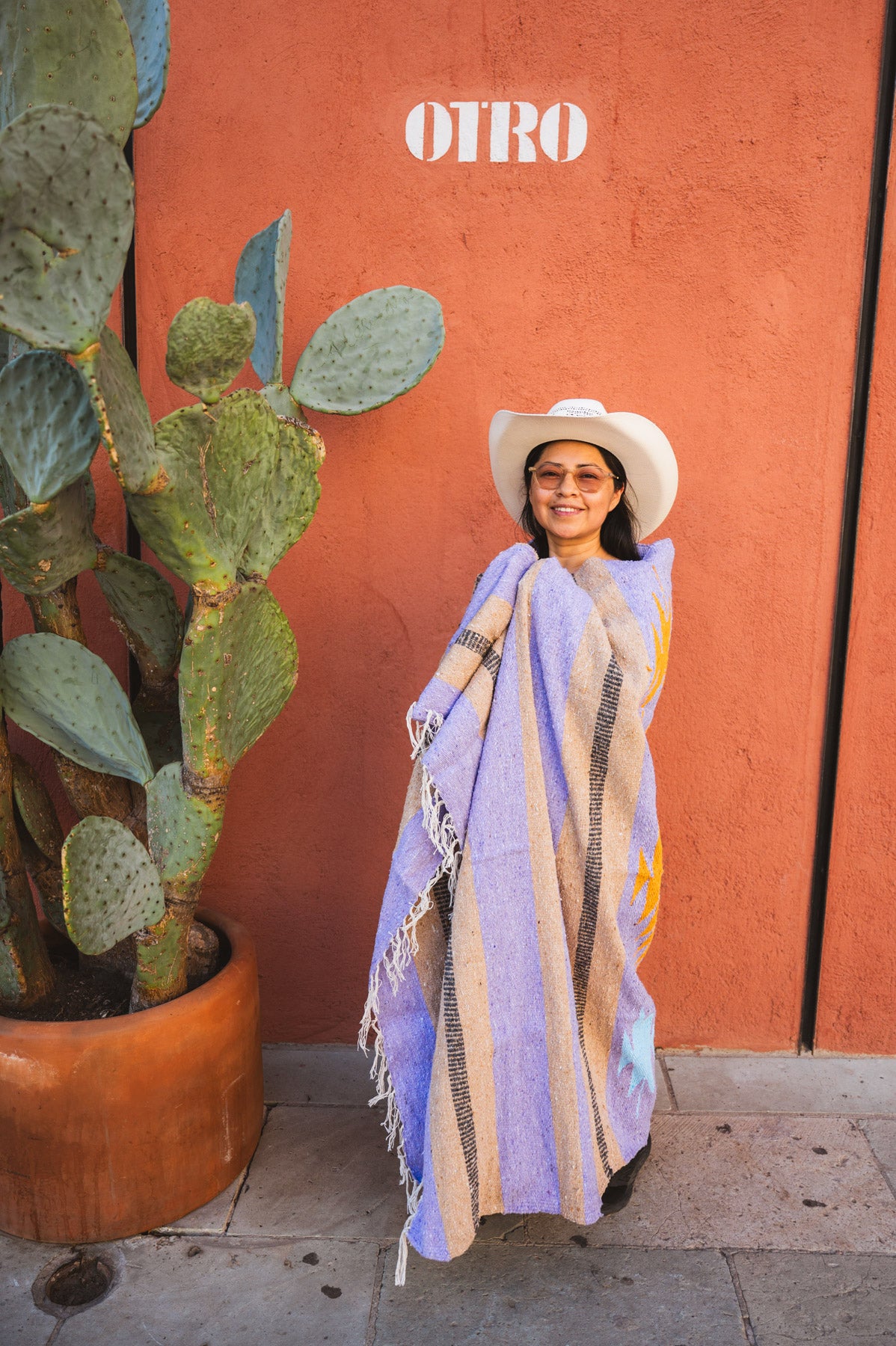 Woman in a purple shawl and white hat standing next to a cactus plant with a red wall in the background.