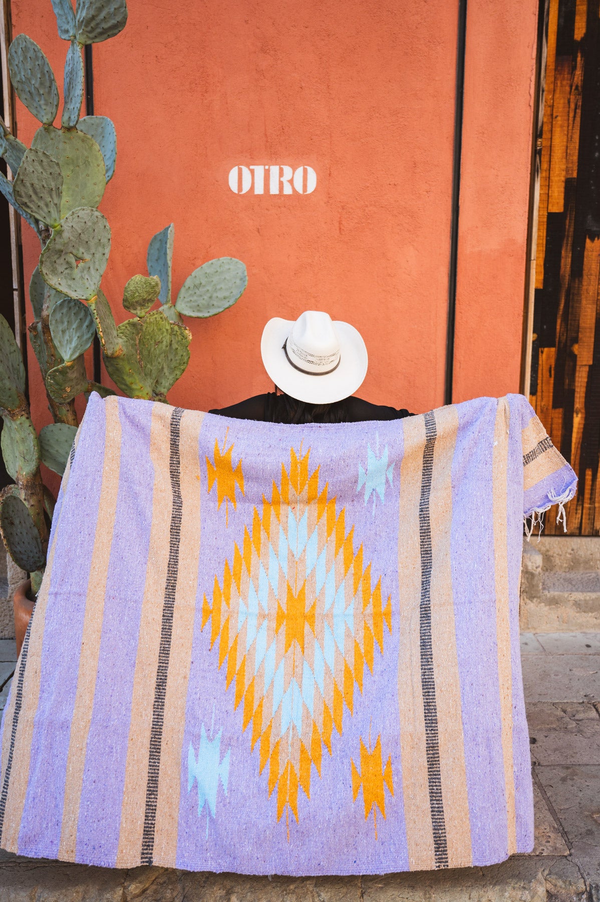 Person draped in a colorful patterned blanket with 'OTRO' branding against an orange wall.