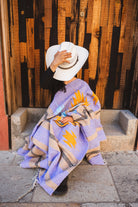 Person sitting on steps wearing a patterned blanket and white hat against a wooden wall.