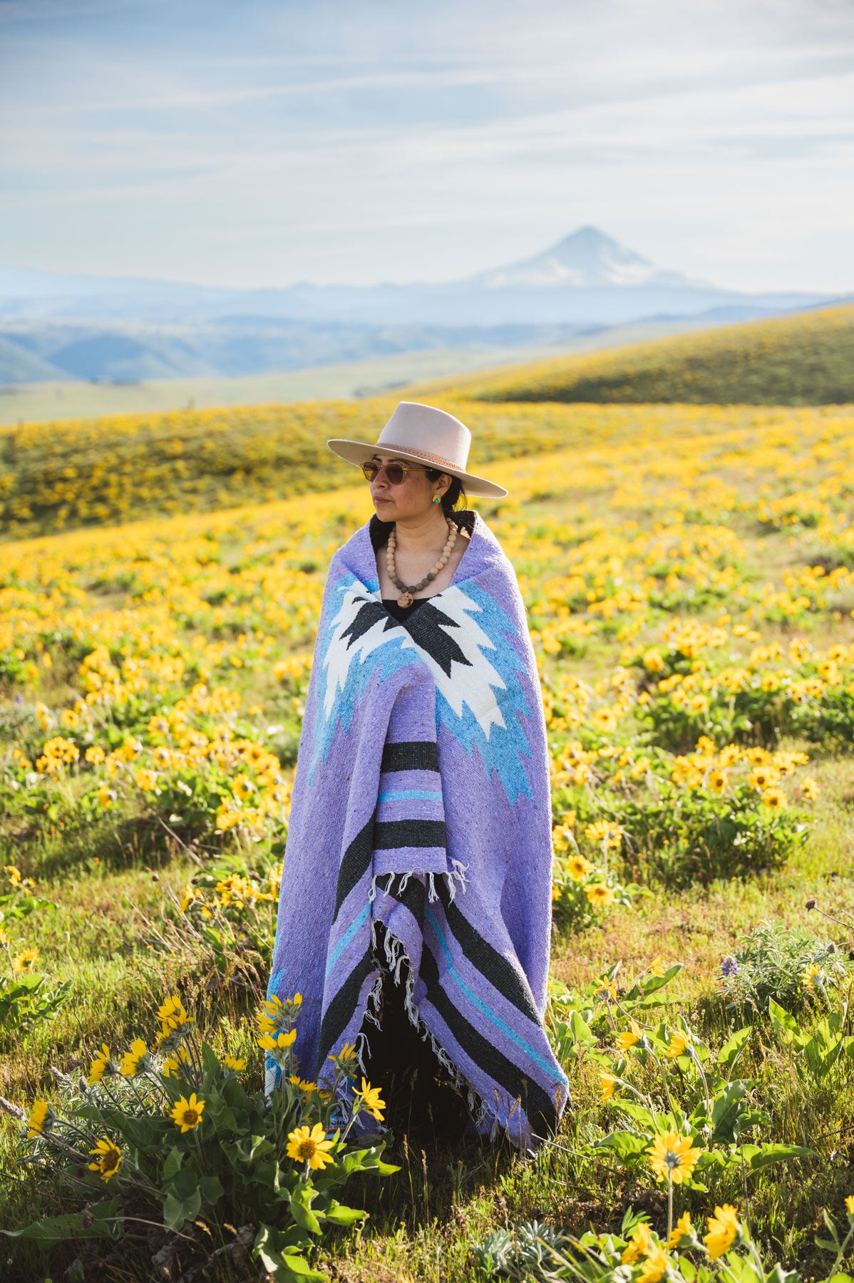 Person wrapped in a colorful blanket standing in a field of yellow flowers with mountains in the background