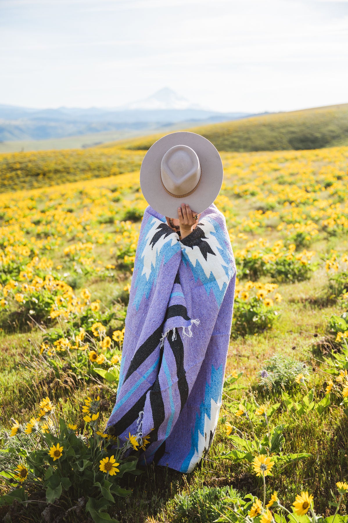 Person wrapped in a colorful blanket standing in a field of yellow flowers with mountains in the background