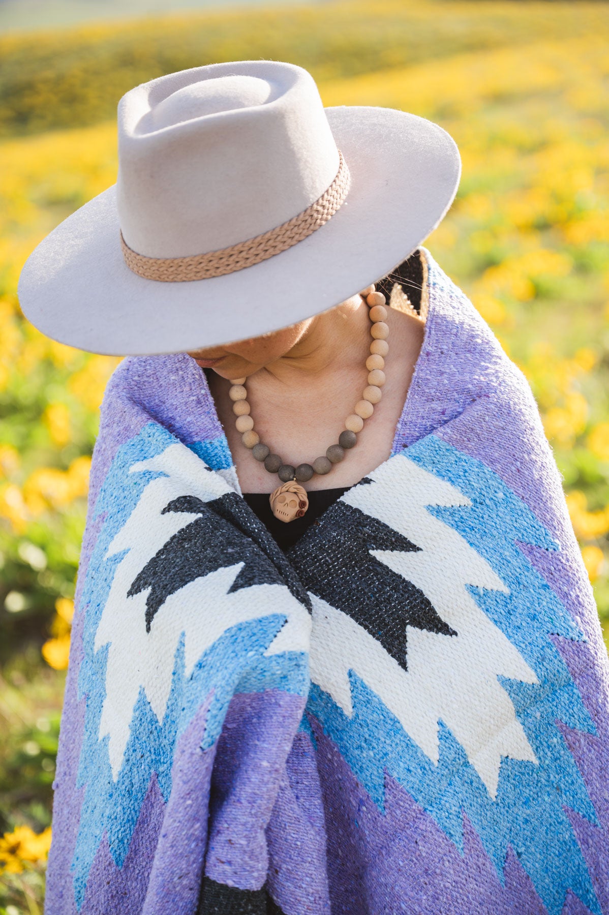 Person wearing a wide-brimmed hat and colorful patterned shawl in a field of yellow flowers.