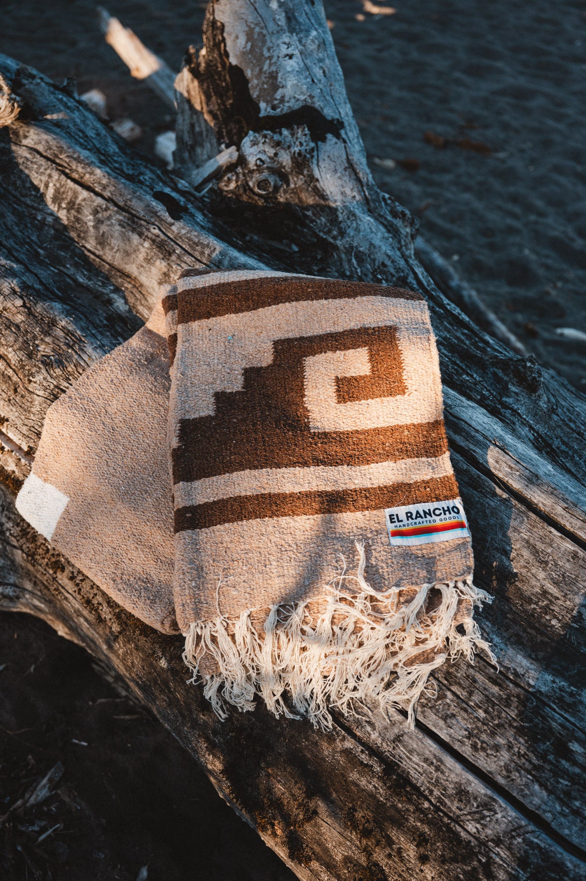 A woven throw blanket with a geometric pattern in brown and cream, draped over a piece of driftwood outdoors. The blanket has fringed edges and a small woven brand label that reads 'EL RANCHO'.