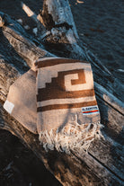 A woven throw blanket with a geometric pattern in brown and cream, draped over a piece of driftwood outdoors. The blanket has fringed edges and a small woven brand label that reads 'EL RANCHO'.
