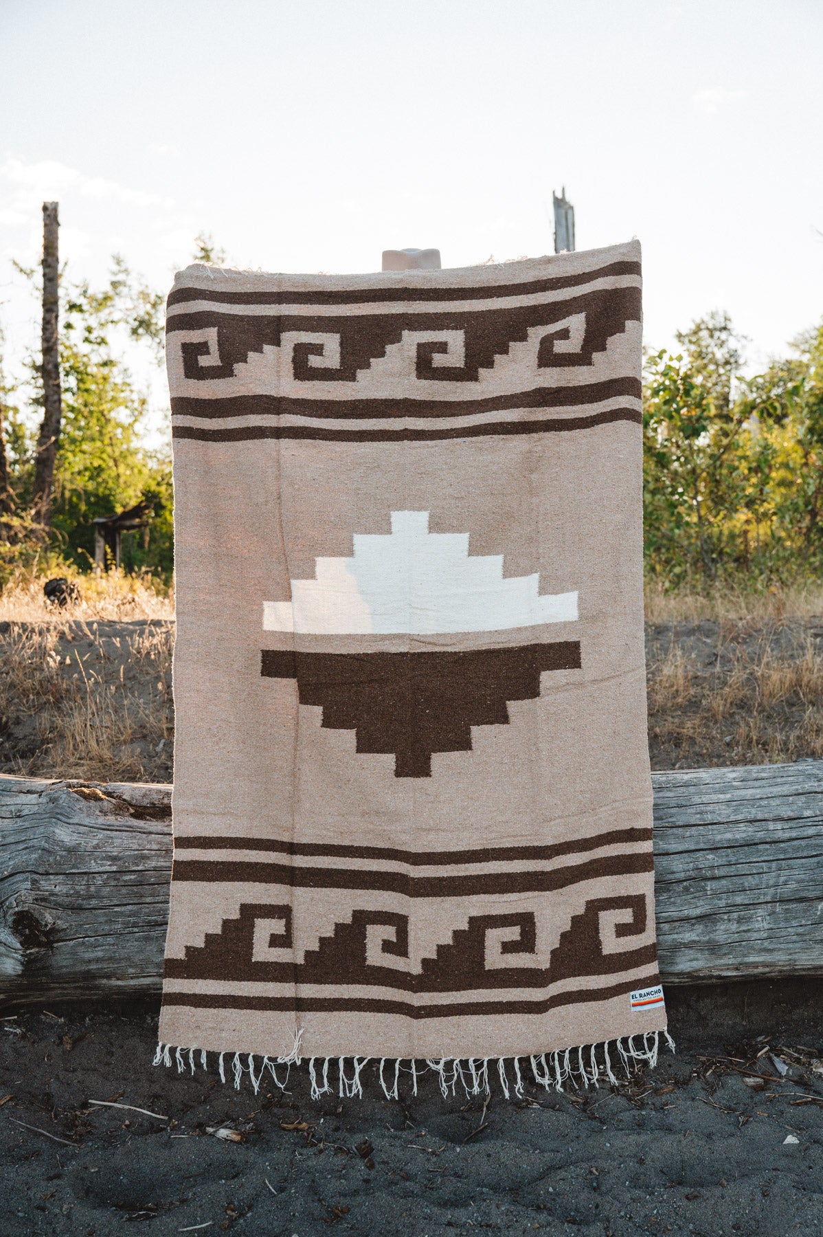 A woven blanket with a Southwestern geometric pattern in beige, brown, and white, hanging outdoors on a rustic wooden stand.