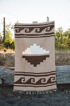 A woven blanket with a Southwestern geometric pattern in beige, brown, and white, hanging outdoors on a rustic wooden stand.