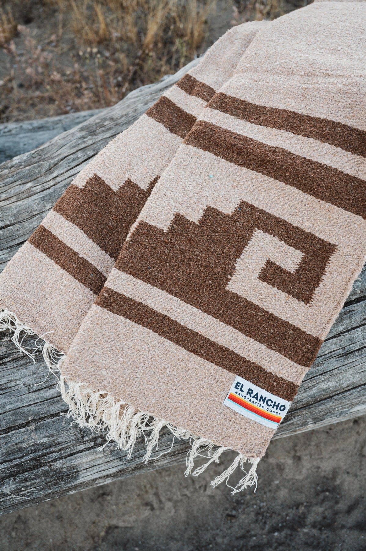 A woven blanket with a brown geometric pattern on a beige background, draped over a weathered wooden log outdoors. The blanket has fringed edges and a small woven label that reads 'EL Rancho Handwoven'.