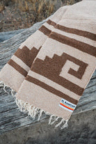A woven blanket with a brown geometric pattern on a beige background, draped over a weathered wooden log outdoors. The blanket has fringed edges and a small woven label that reads 'EL Rancho Handwoven'.