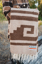 A person outdoors holds up a woven blanket with a brown and beige geometric pattern and fringe. The blanket has a visible El Rancho Handwoven label near the bottom right corner.