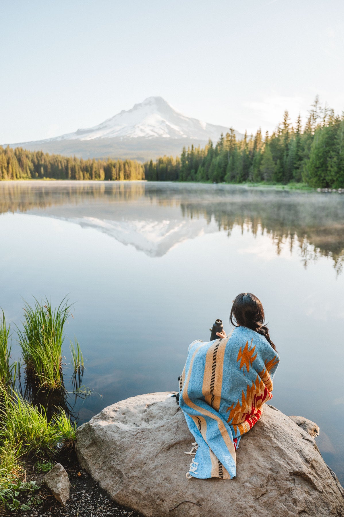 Girl wrapped in blue handwoven mexican blanket sitting on rock in front of Trillium lake looking at Mt. Hood