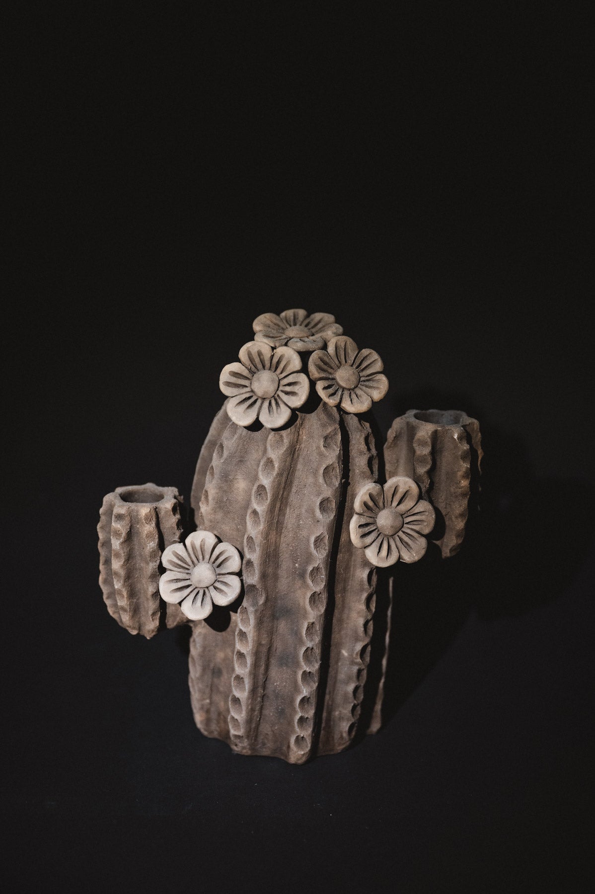 A sculptural cactus-shaped ceramic vase with textured vertical ridges and five raised flower motifs, shown on a solid black background.