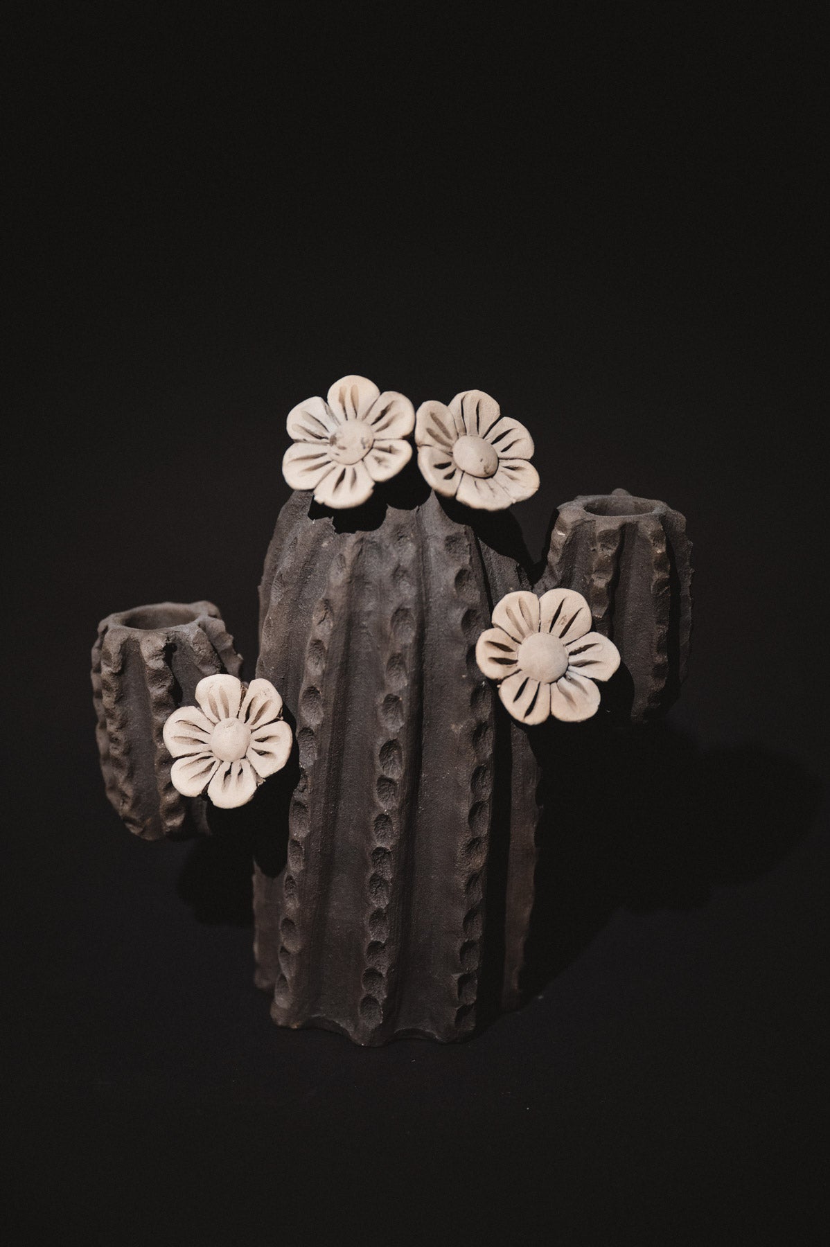 A sculptural dark gray vase or planter with a ribbed, textured cylindrical body and three short side arms, decorated with four off-white flower shapes on top and sides, photographed against a solid black background.