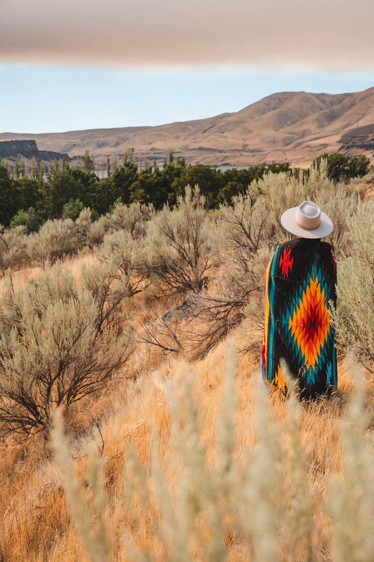 A person standing outdoors in a dry grassy landscape, viewed from behind, wearing a wide-brim white hat and a colorful geometric patterned blanket or poncho with red, orange, yellow, green, and blue motifs.