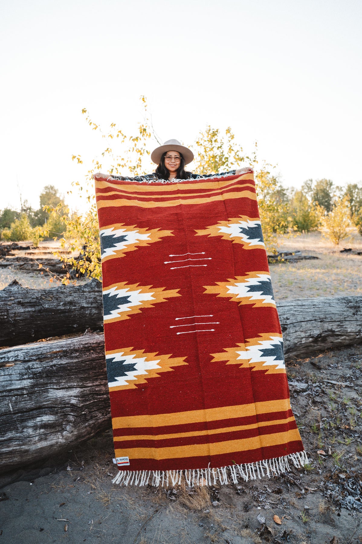Person holding a large relampago patterned mexican blanket outdoors with trees and logs in the background