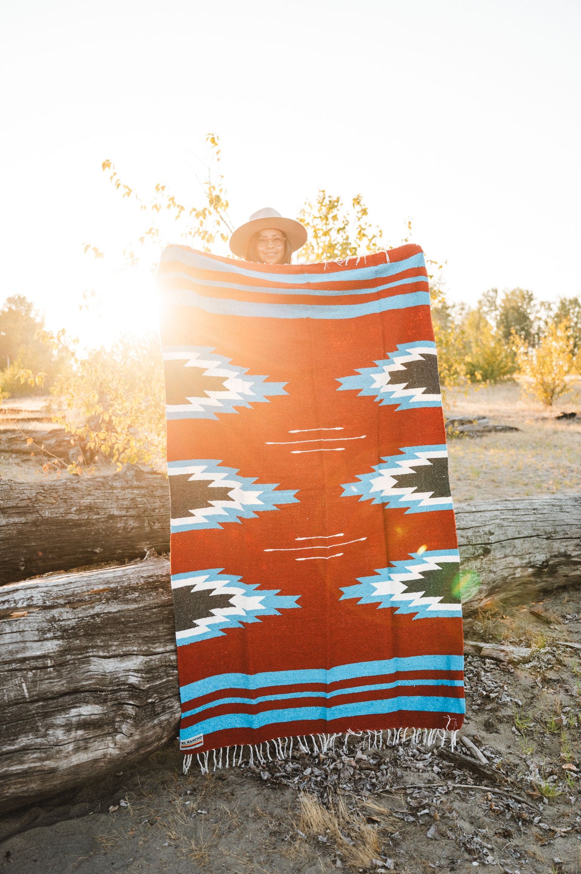 Person holding a large relampago patterned mexican blanket outdoors with trees and logs in the background