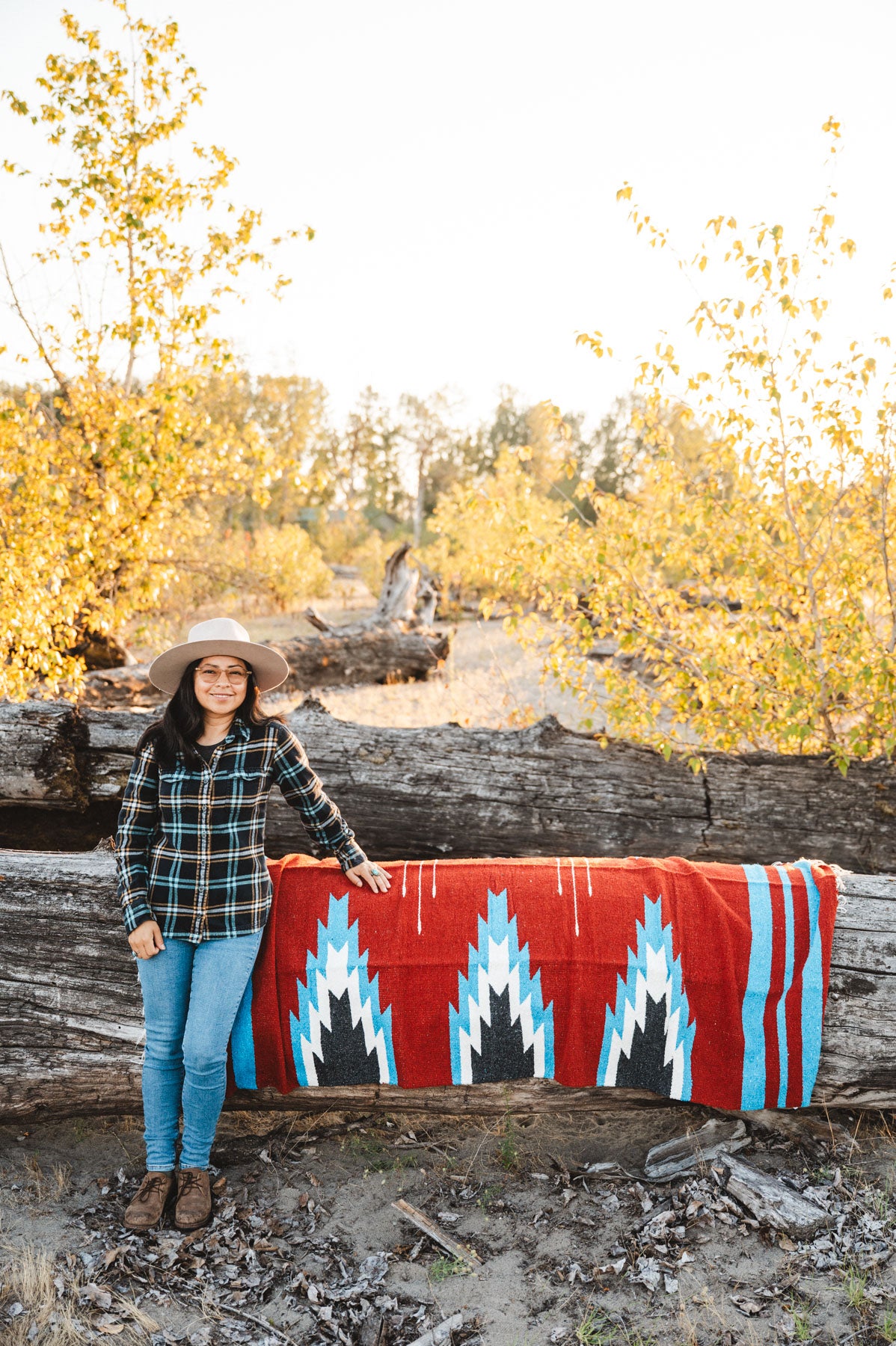 Person sitting next to a large relampago patterned mexican blanket outdoors with trees and logs in the background