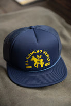 A navy blue trucker hat with a yellow embroidered logo reading 'EL RANCHO SUPPLY' and a donkey graphic, resting on a textured brown surface.