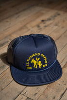 A navy blue trucker hat with yellow embroidered text reading 'EL RANCHO SUPPLY' and a donkey logo, resting on a rustic wooden floor.