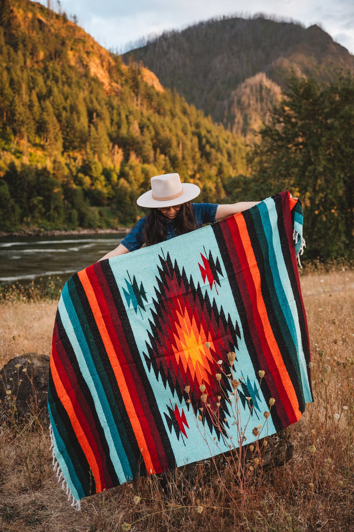 Woman holding teal handwoven diamond mexican blanket in front of mountain at sunset