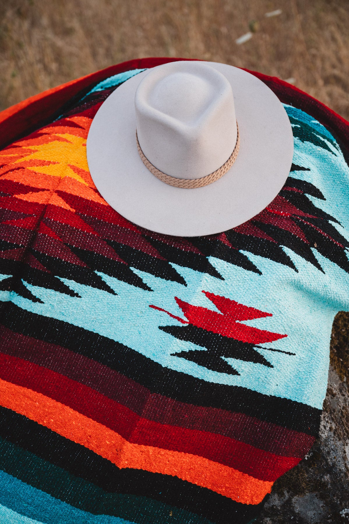 A white cowboy hat with a tan rope band rests on a colorful woven blanket featuring geometric patterns in turquoise, black, red, orange, and dark blue.