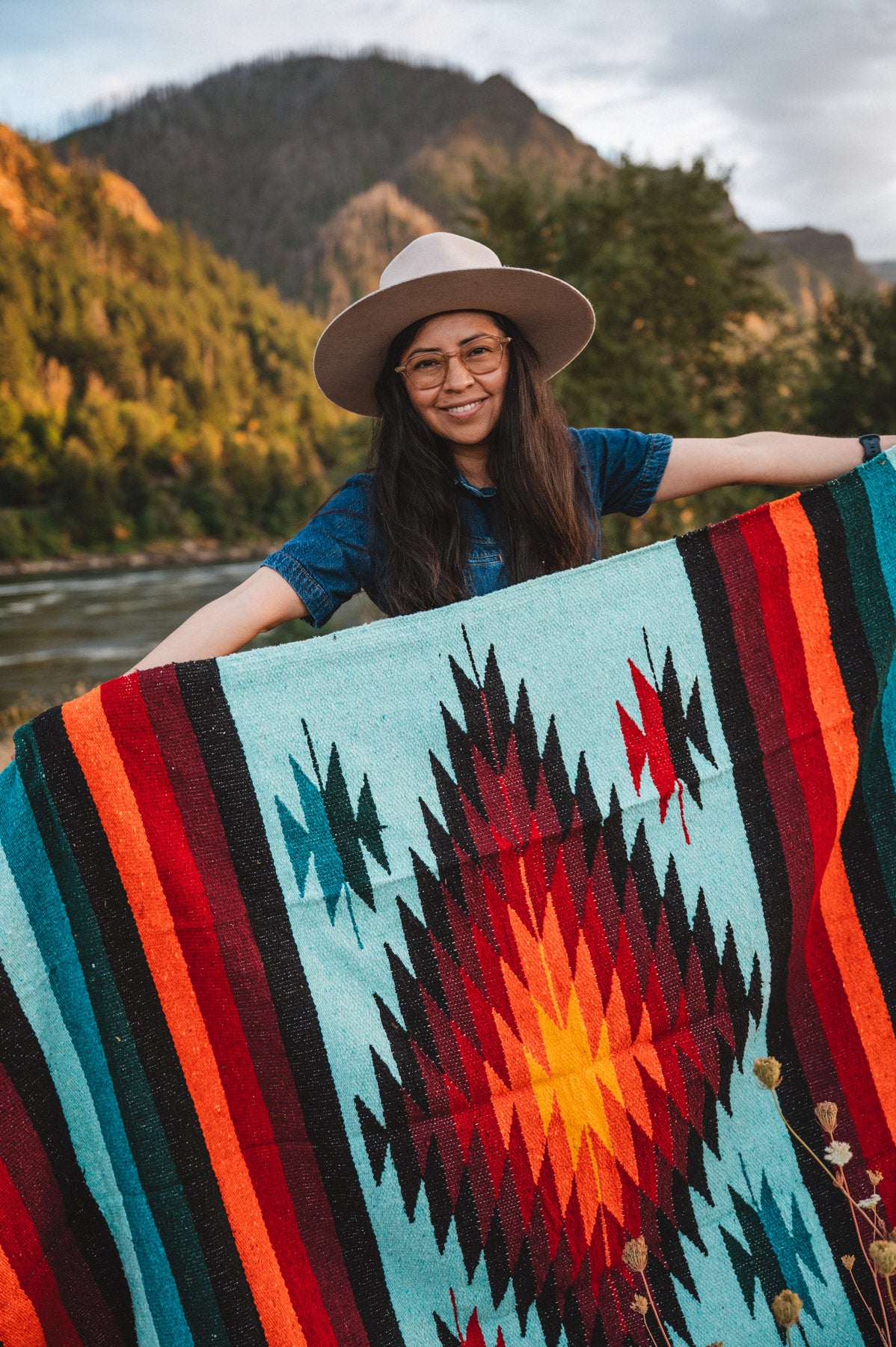 Woman holding teal handwoven diamond mexican blanket in front of mountain at sunset