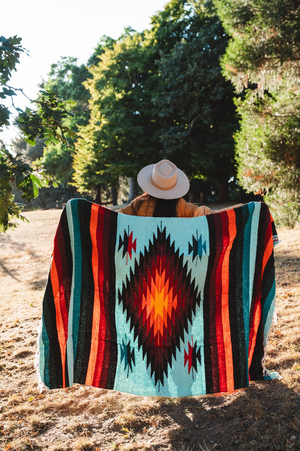 A person sitting outdoors in a wooded area, viewed from behind, wrapped in a large Southwestern-style woven blanket. The blanket features a turquoise background with a large red, orange, and black diamond motif, flanked by vertical stripes of red, orange, black, and teal.