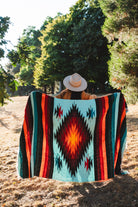 A person sitting outdoors in a wooded area, viewed from behind, wrapped in a large Southwestern-style woven blanket. The blanket features a turquoise background with a large red, orange, and black diamond motif, flanked by vertical stripes of red, orange, black, and teal.