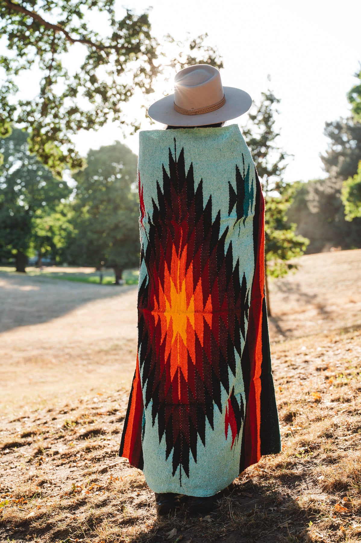 A person standing outdoors in a park wearing a wide-brimmed straw hat and a long woven blanket draped over their body, showing a Southwestern-style geometric pattern in turquoise, black, red, and orange.