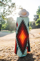 A person standing outdoors in a park, viewed from behind, wearing a wide-brimmed light beige hat and a long woven blanket or wrap with a bold Southwestern geometric pattern in turquoise, black, red, orange, and yellow.