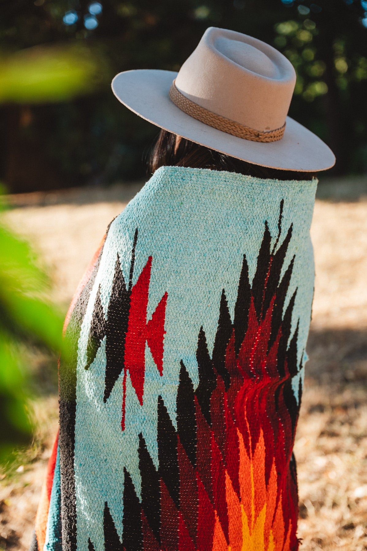 A person outdoors wearing a wide-brim beige hat and a light blue woven blanket or poncho with bold black, red, and orange geometric patterns, shown from the back.