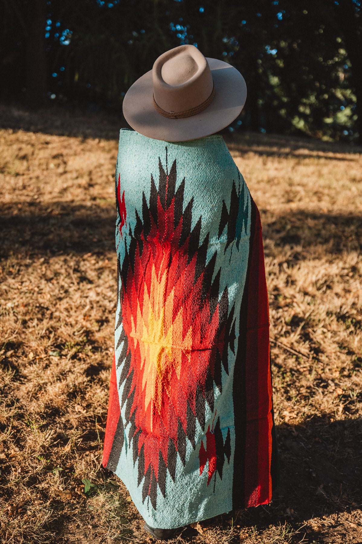 A person standing outdoors in a dry grassy field, viewed from behind, wearing a wide-brim beige hat and a large woven blanket or poncho with a Southwestern-style geometric pattern in turquoise, red, orange, black, and white.