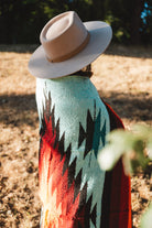 A person outdoors wearing a wide-brim beige hat and a colorful woven blanket or poncho with a Southwestern-style geometric pattern in turquoise, black, red, and orange.