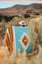 A person wearing a wide-brim hat sits outdoors in a desert landscape, wrapped in a large woven blanket with turquoise, mustard, white, and brown geometric patterns.
