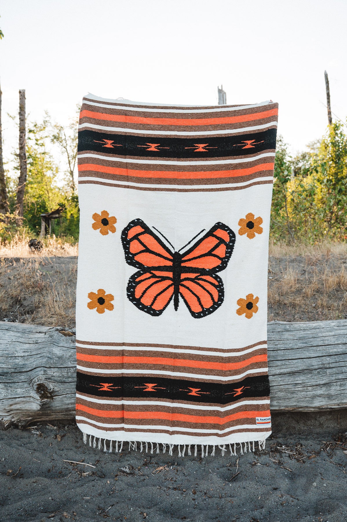A woven blanket with a large orange and black butterfly design, surrounded by small orange flowers and horizontal striped bands, draped over a log outdoors.