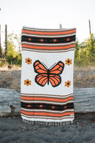 A woven blanket with a large orange and black butterfly design, surrounded by small orange flowers and horizontal striped bands, draped over a log outdoors.