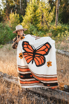 A person outdoors in a wooded, grassy setting holds up a large woven blanket featuring a prominent orange and black butterfly design on a cream background, with orange and brown floral and striped accents.