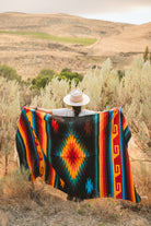 A person standing outdoors in a dry grassy landscape holds up a large woven blanket with a bold Southwestern geometric pattern in black, orange, red, yellow, and blue.