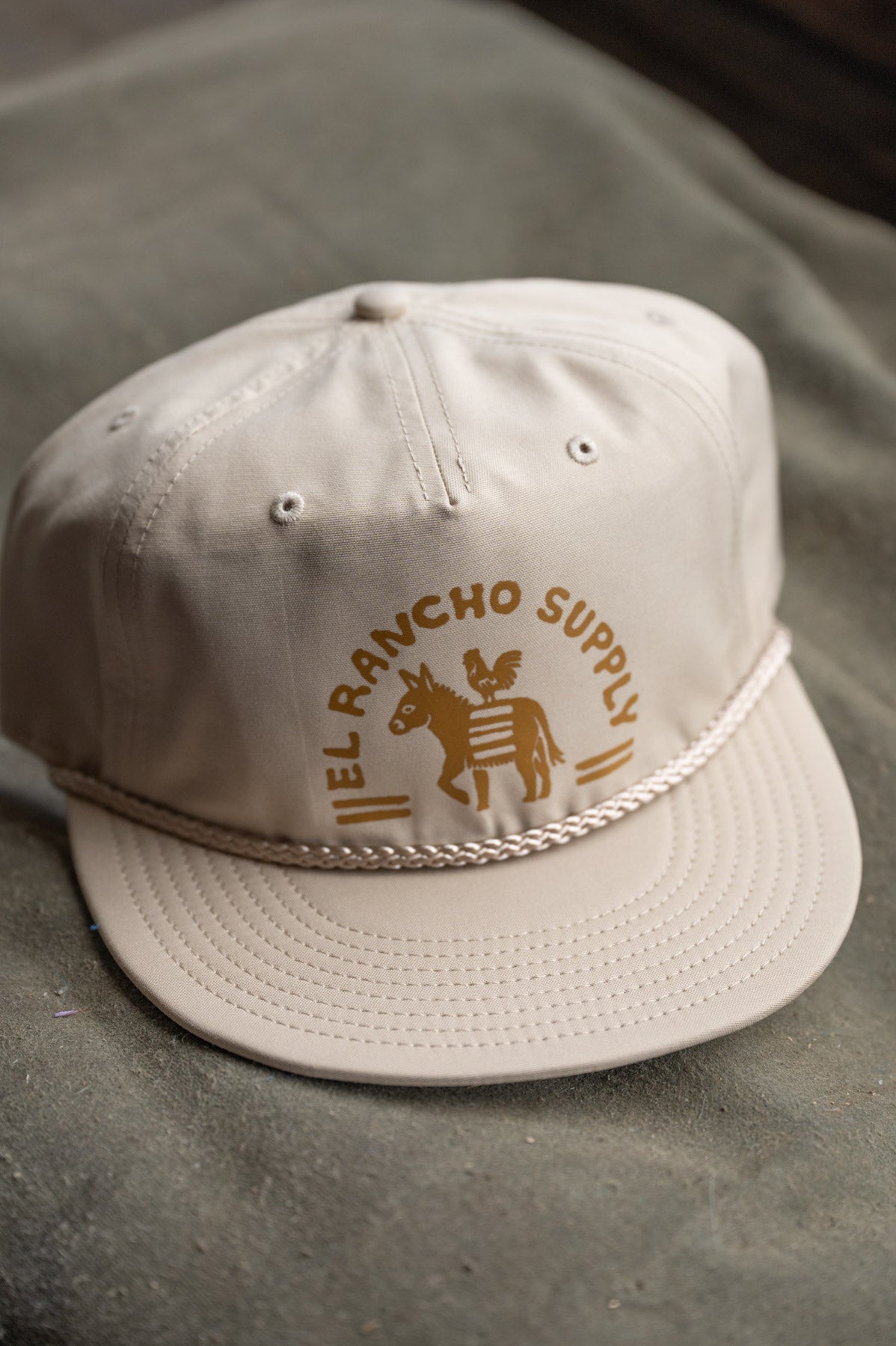 A light beige baseball cap with a brown embroidered logo reading 'EL RANCHO SUPPLY' and a rope detail around the brim, resting on a textured gray surface.