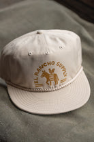 A light beige baseball cap with a brown embroidered logo reading 'EL RANCHO SUPPLY' and a rope detail around the brim, resting on a textured gray surface.